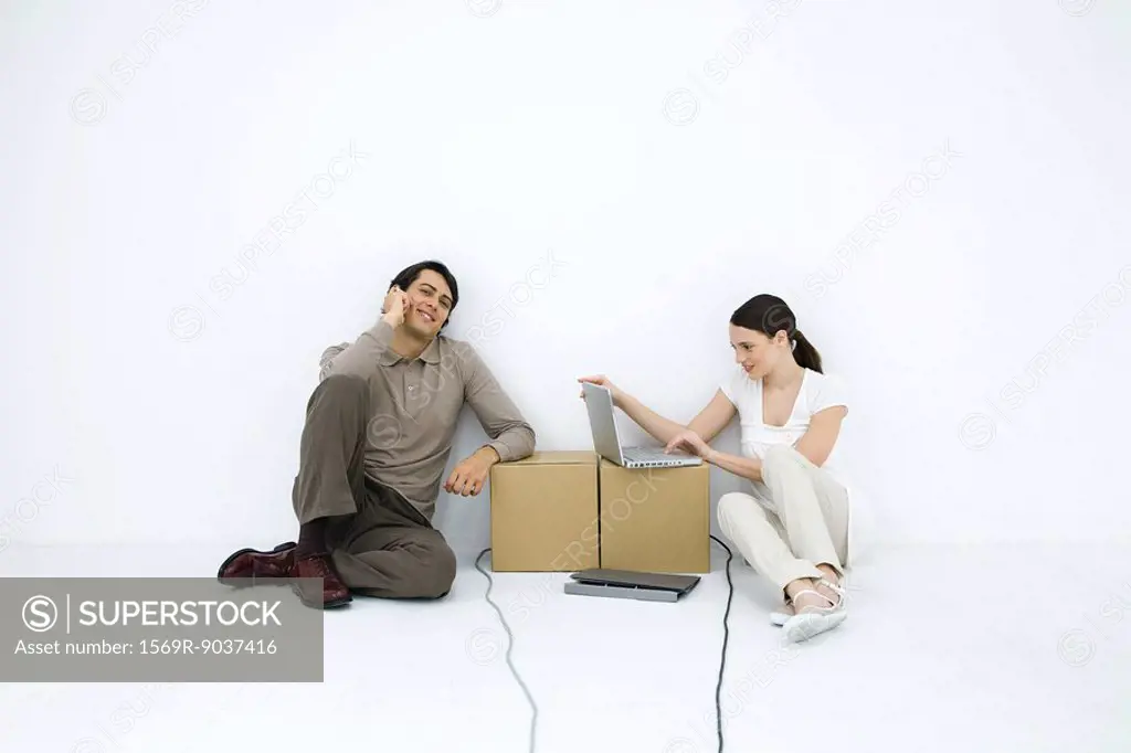 Man talking on phone, sitting on floor next to woman using laptop computer at cardboard box desk