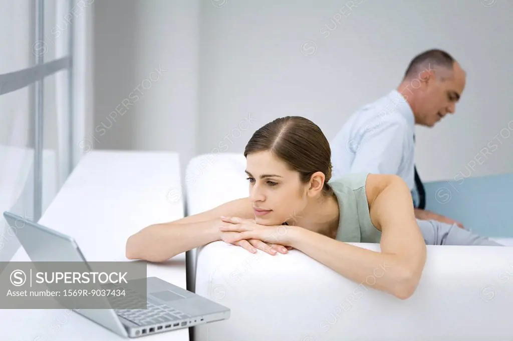 Young woman relaxing on sofa, using laptop computer, businessman reading in background
