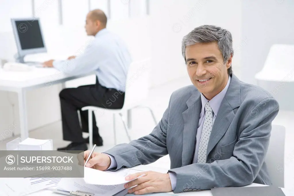 Businessman working at desk, smiling at camera, colleague in background