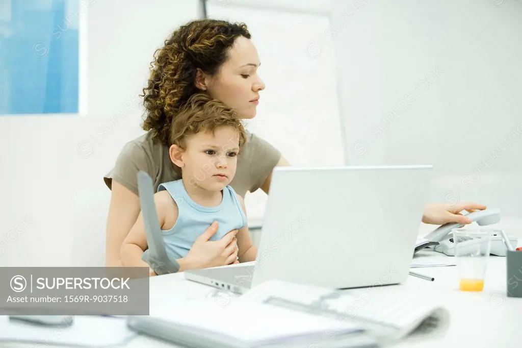 Woman sitting at desk with toddler on her lap, hanging up landline phone