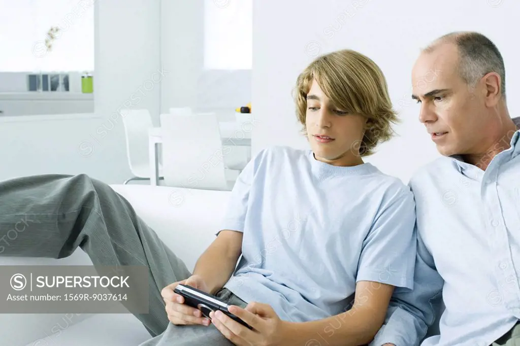 Teen boy playing handheld video game, father watching over his shoulder
