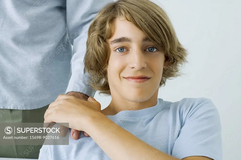 Teen boy holding father´s hand on shoulder, smiling at camera, cropped view