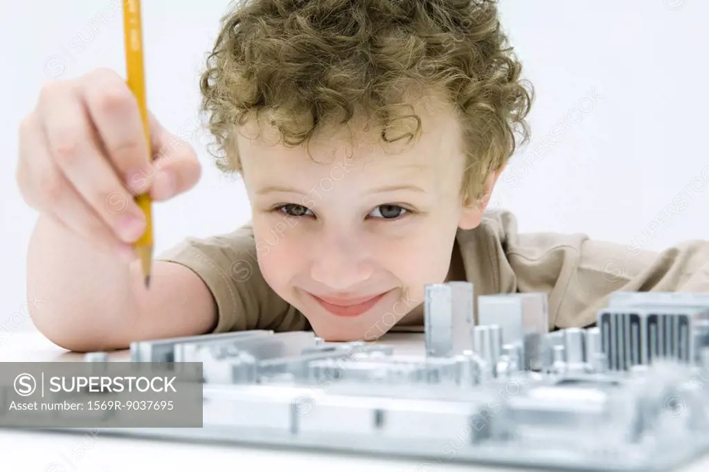 Little boy holding pencil over computer motherboard, smiling at camera