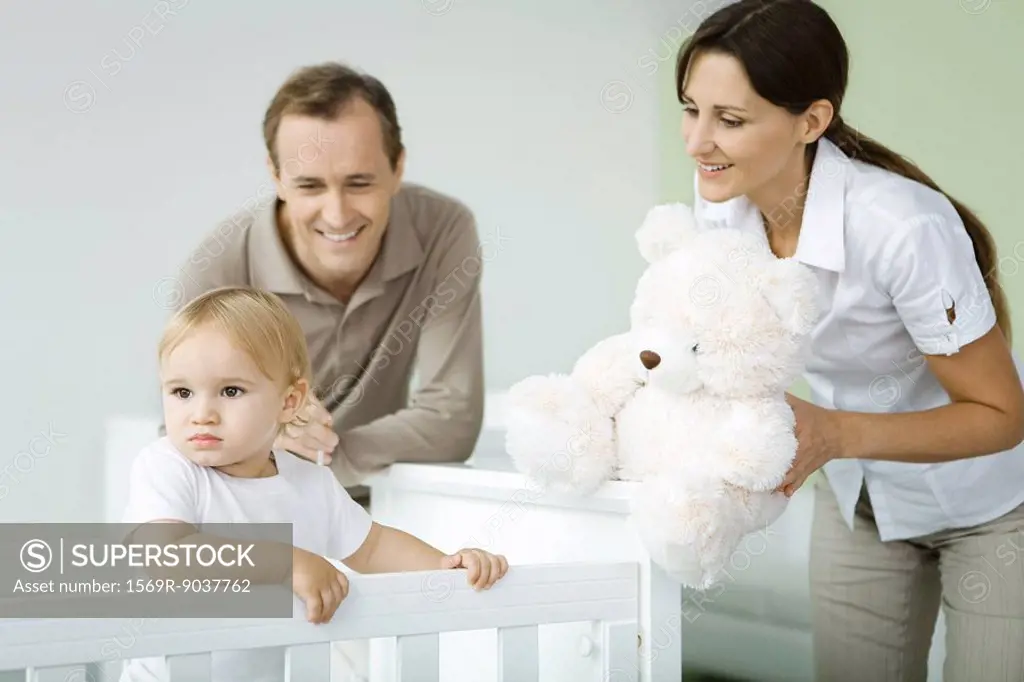 Parents standing beside toddler in crib, mother holding teddy bear, toddler looking away