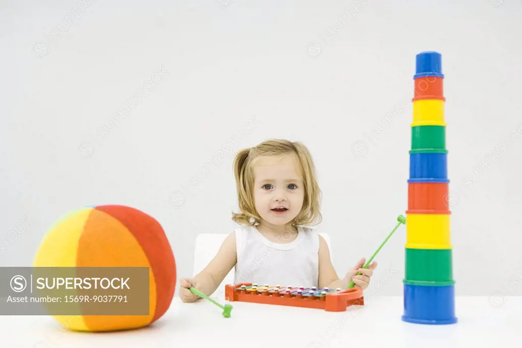 Toddler girl sitting with toys, playing xylophone, smiling at camera