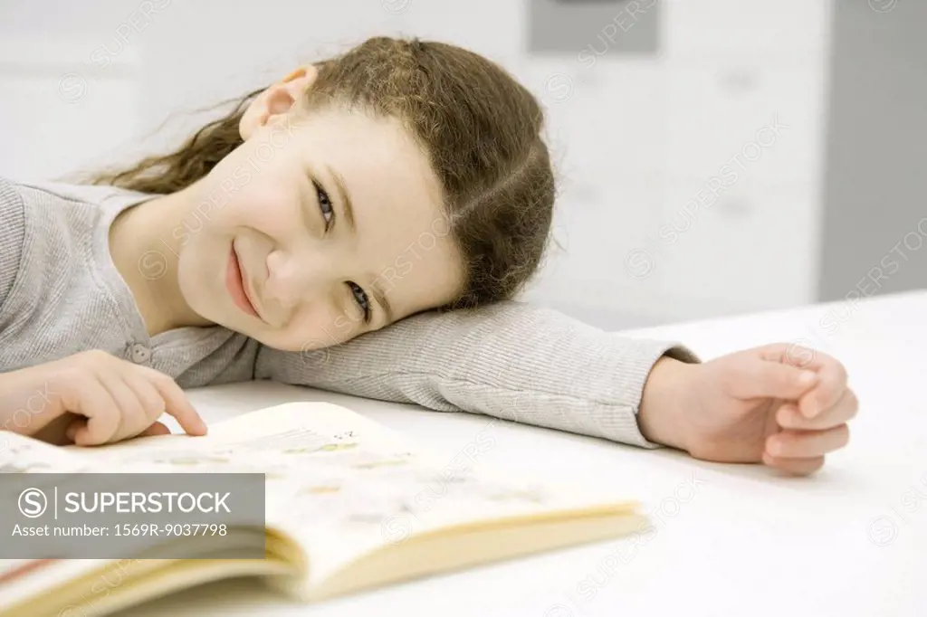 Girl sitting at table with book, resting head on arm, smiling at camera