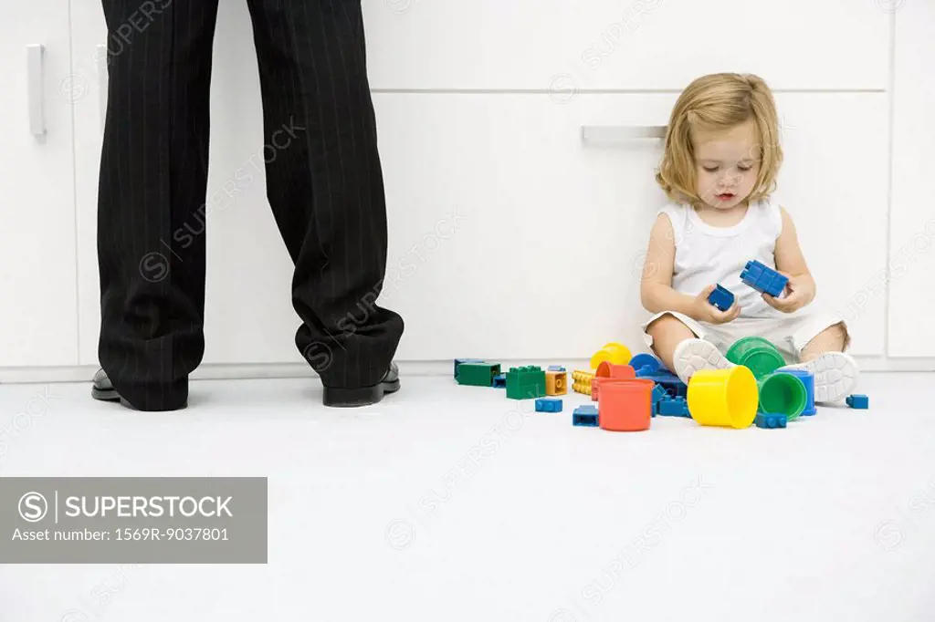 Toddler girl sitting on the ground beside father´s legs, playing with toys
