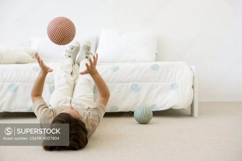 Boy lying on the ground in bedroom, throwing ball in the air