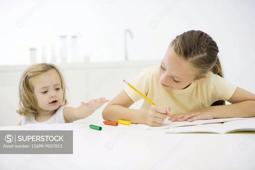 Girl doing homework at table, younger sister reaching for crayons