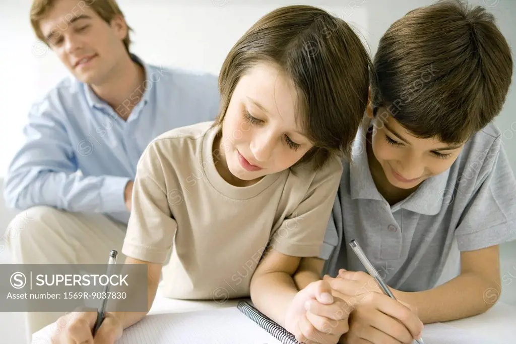 Two boys writing in notebook together, father watching in background