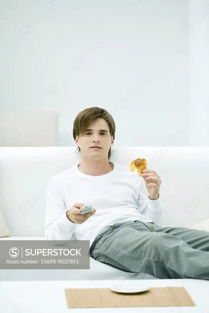 Young man sitting on sofa, holding remote control and croissant