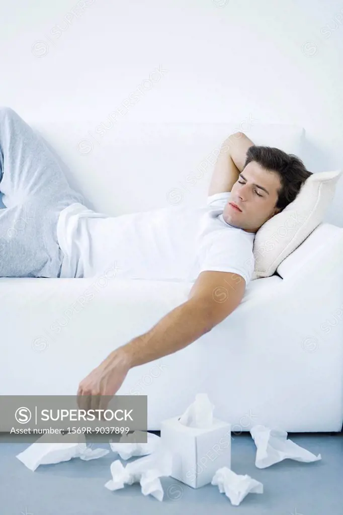 Man lying on sofa, discarded tissues on the floor beside him