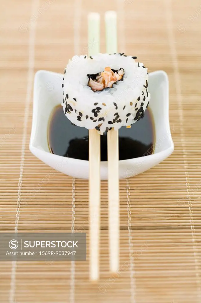 Maki sushi balanced on chopsticks over dish of soy sauce