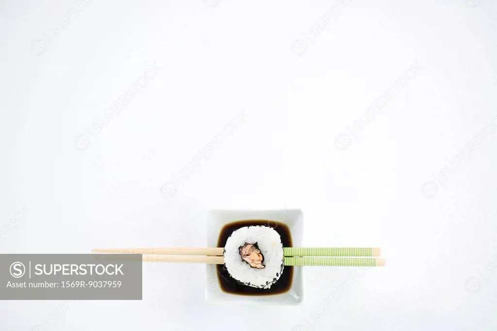 Single piece of maki sushi resting on chopsticks over dish of soy sauce, overhead view