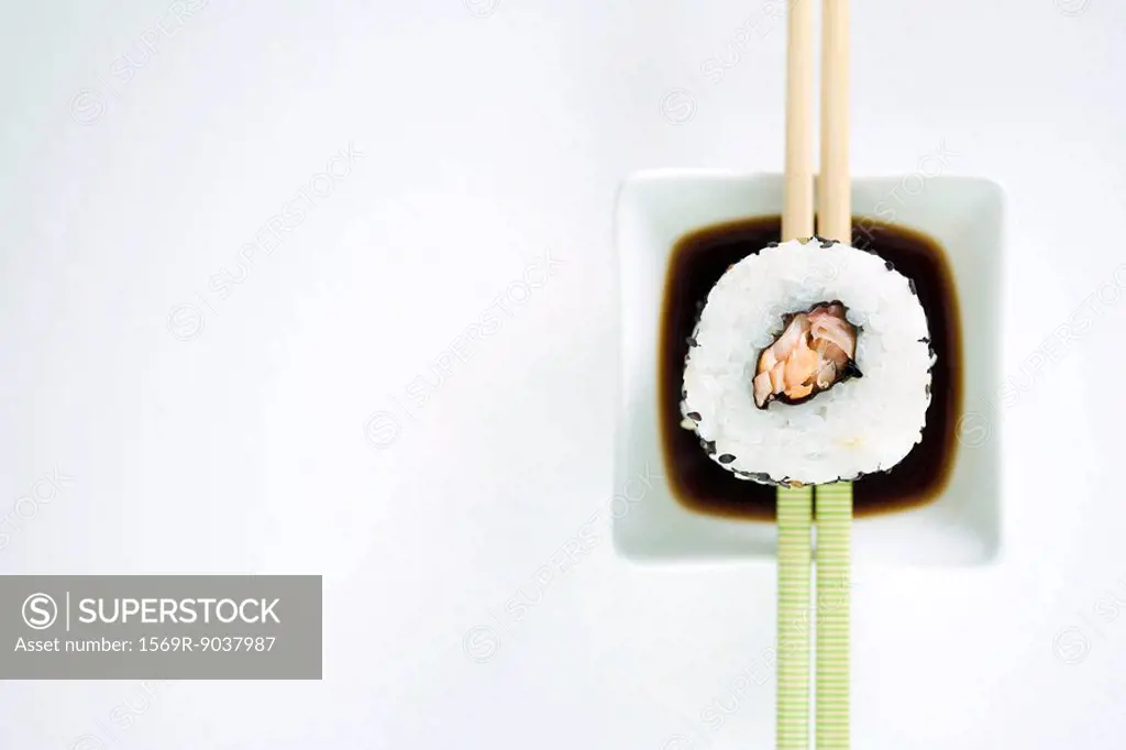 Single piece of maki sushi resting on chopsticks over sauce, overhead view