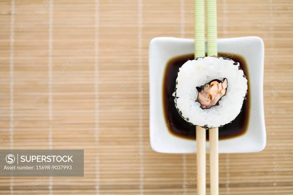 Single piece of maki sushi resting on chopsticks over sauce, overhead view