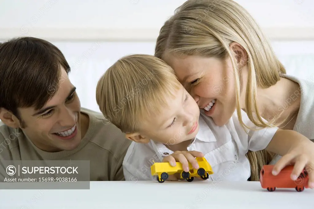 Little boy sitting with parents, playing with toy trucks, mother nuzzling his cheek