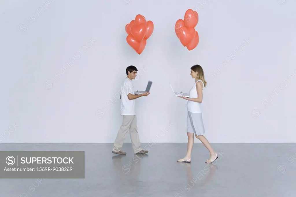 Young man and woman walking toward each other, both carrying laptop computers and heart balloons