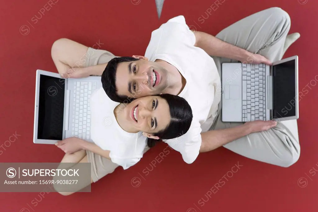 Couple sitting back to back on the ground, holding laptop computers, smiling up at camera