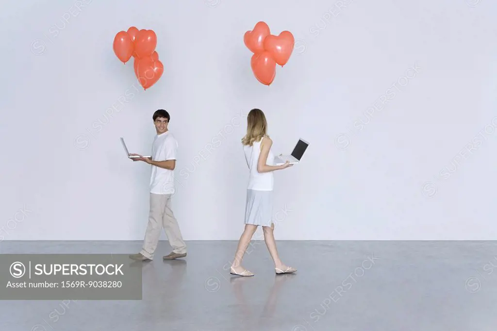 Man and woman walking past each other, both carrying laptop computers and heart balloons