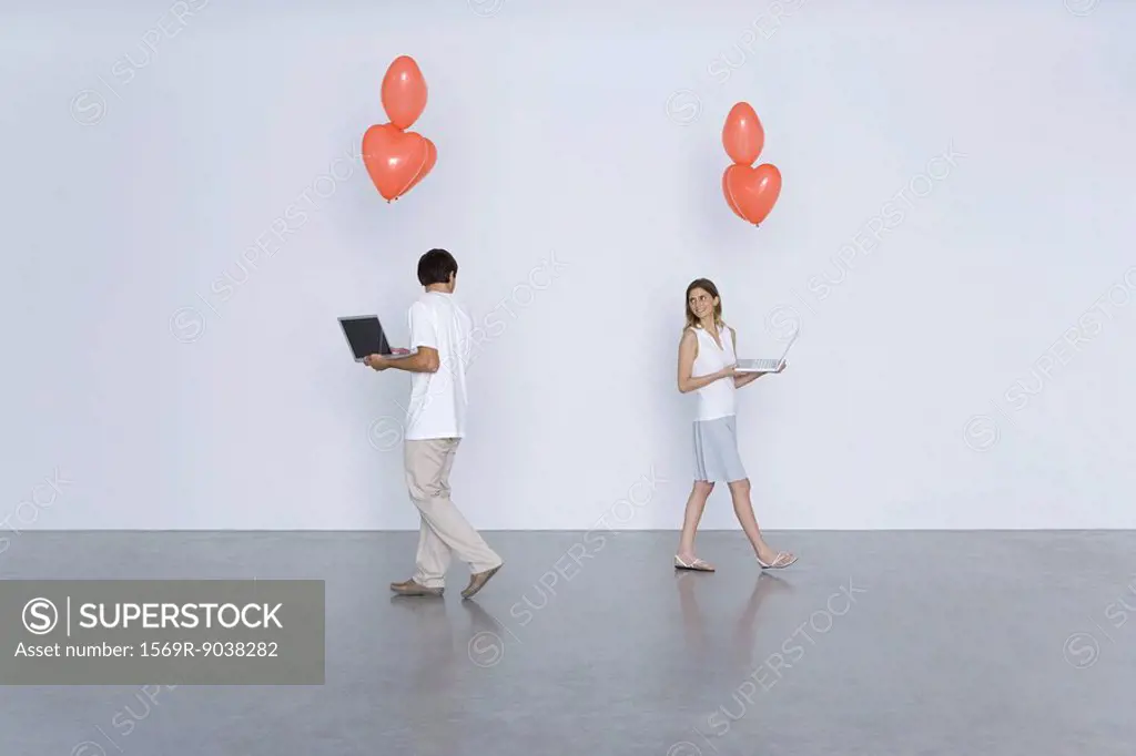 Man and woman carrying laptop computers and heart balloons, smiling over their shoulders at each other
