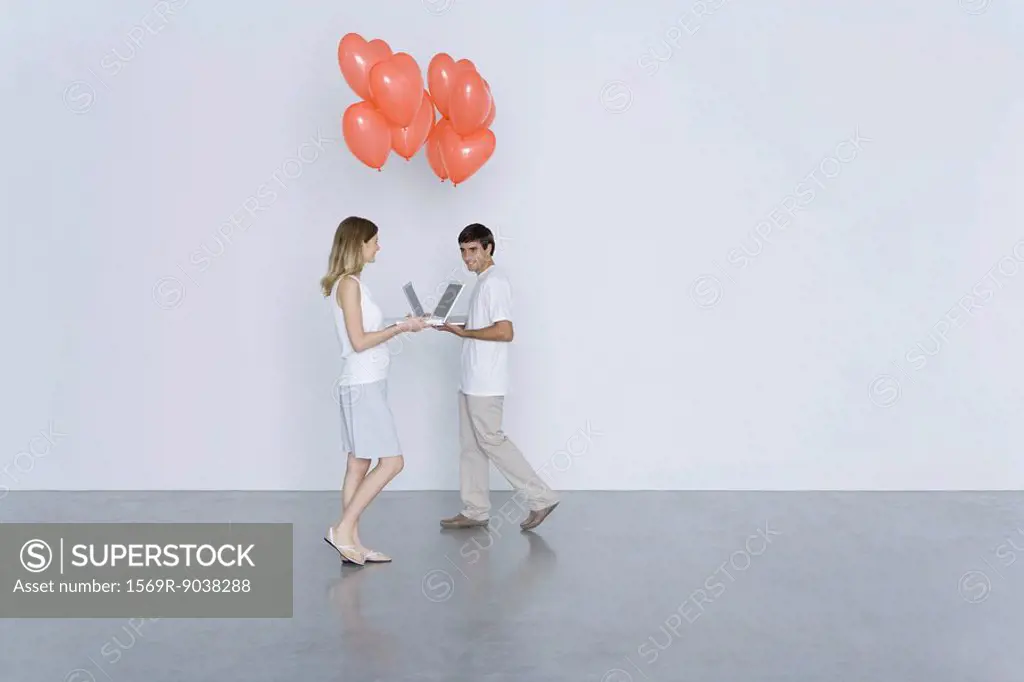Man and woman walking toward each other with laptop computers and heart balloons, smiling