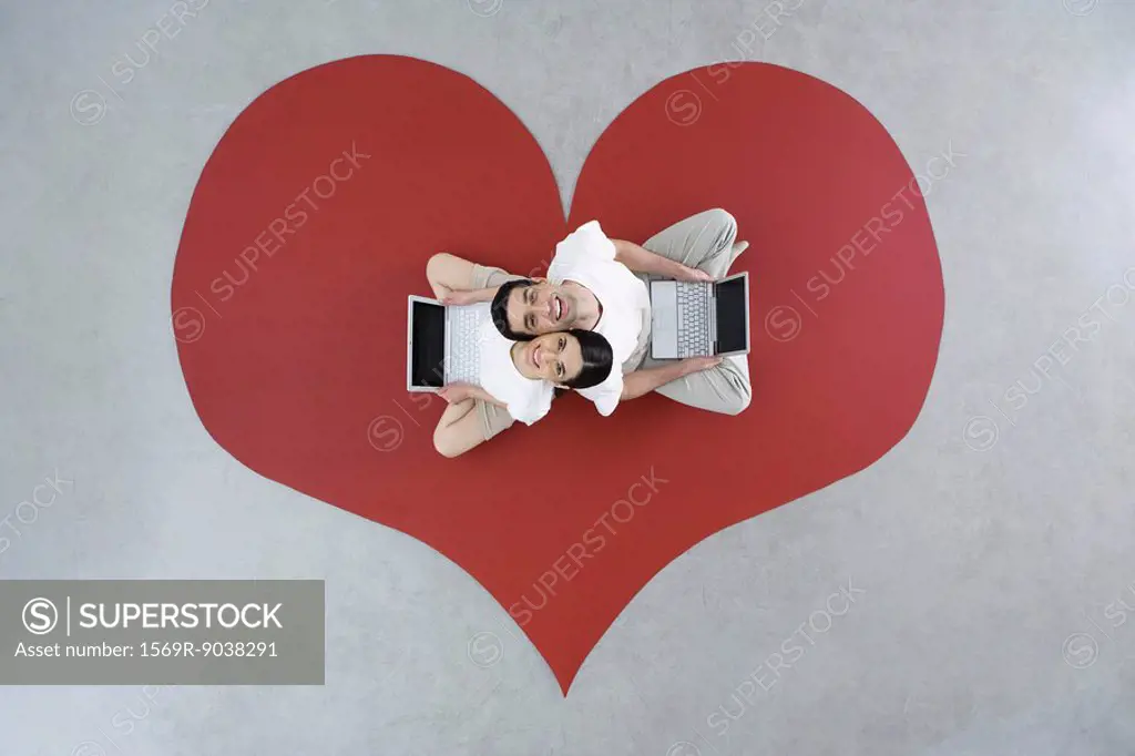 Couple sitting back to back on large heart, holding laptop computers, smiling up at camera