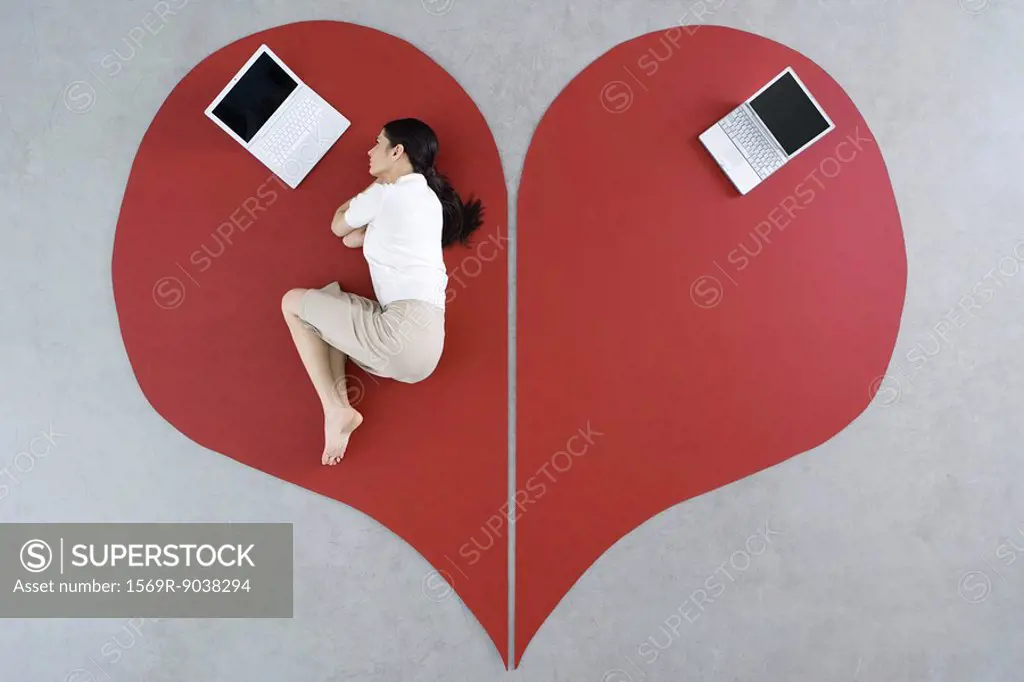 Woman lying on the ground with laptop on large broken heart, abandoned laptop behind her