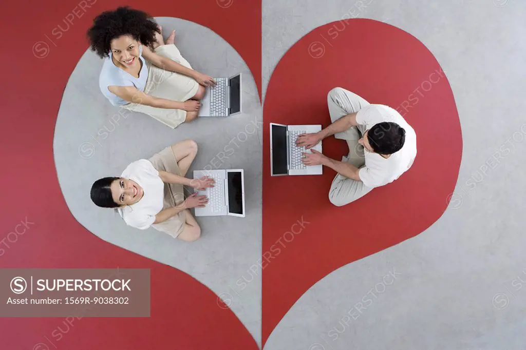 Two women and one man sitting on large heart, using laptop computers, women smiling up at camera