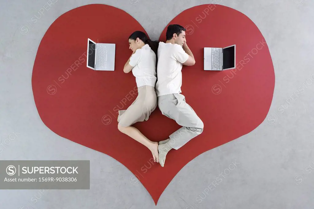Man and woman lying back to back on large heart, both looking at laptop computers