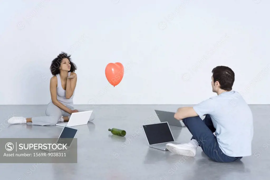 Man and woman seated with laptops, woman blowing man a kiss, bottle and heart balloon between them