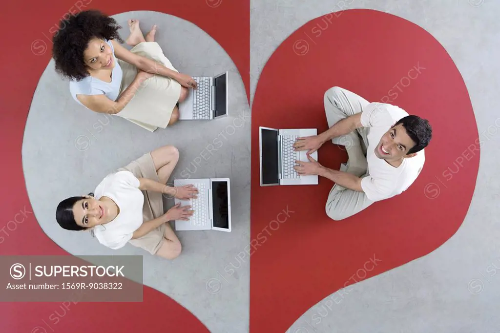 Two women and one man sitting on large heart, using laptop computers, smiling up at camera