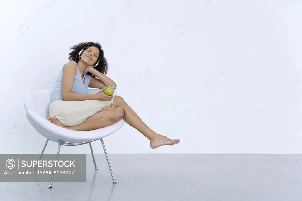 Woman sitting in chair, listening to headphones connected to apple, smiling