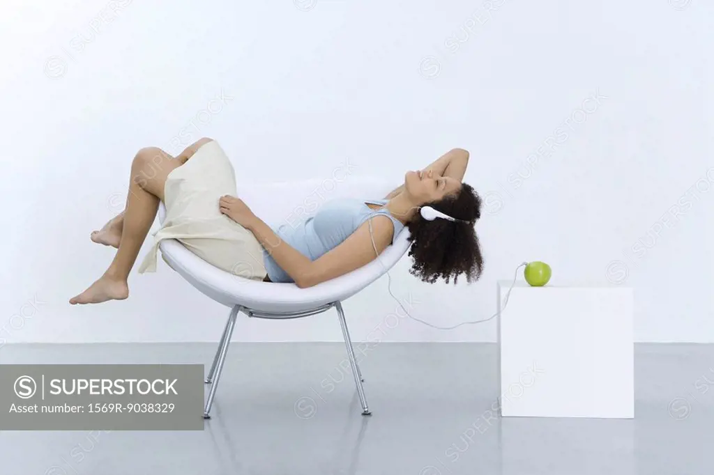 Woman reclining in chair, listening to headphones connected to apple, side view