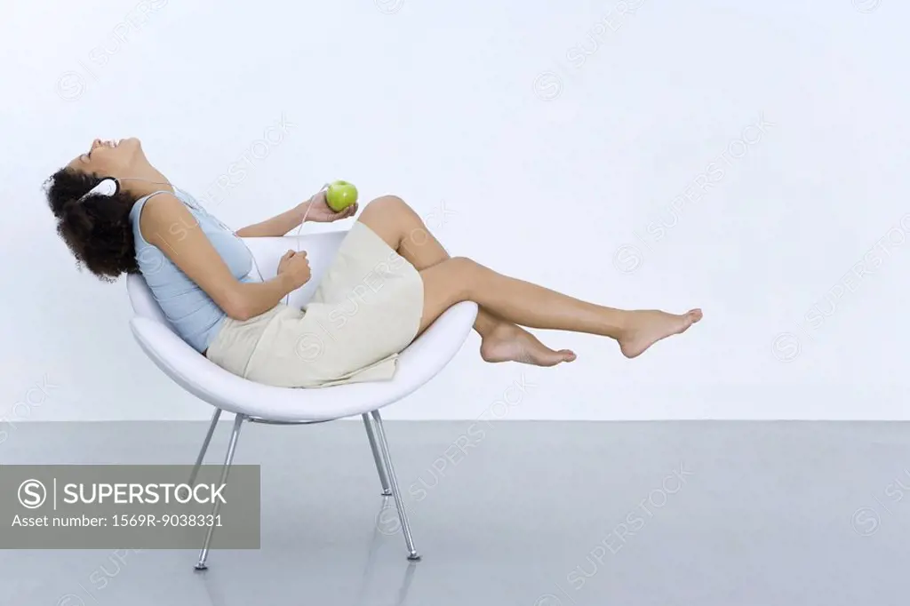 Woman reclining in chair, listening to headphones connected to apple, side view