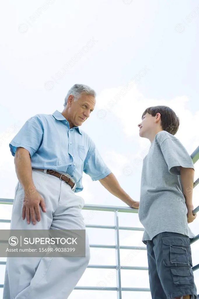 Grandfather and grandson standing by railing, looking at each other, low angle view