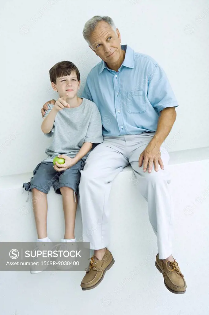 Grandfather and grandson sitting together on ledge, looking away, boy pointing