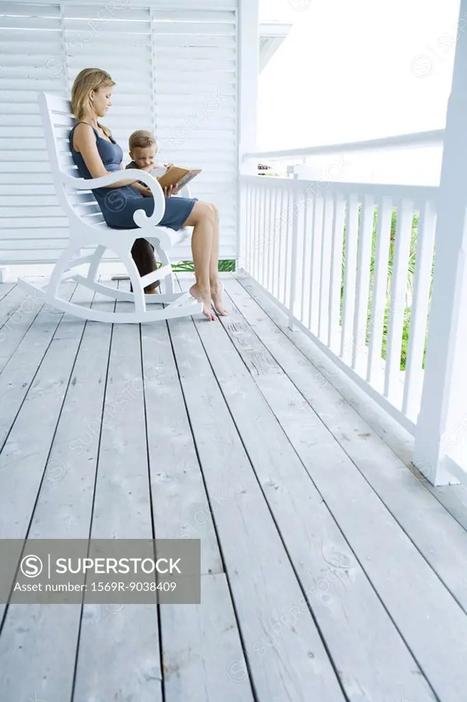 Woman sitting in rocking chair on porch, reading story to little boy standing at her side