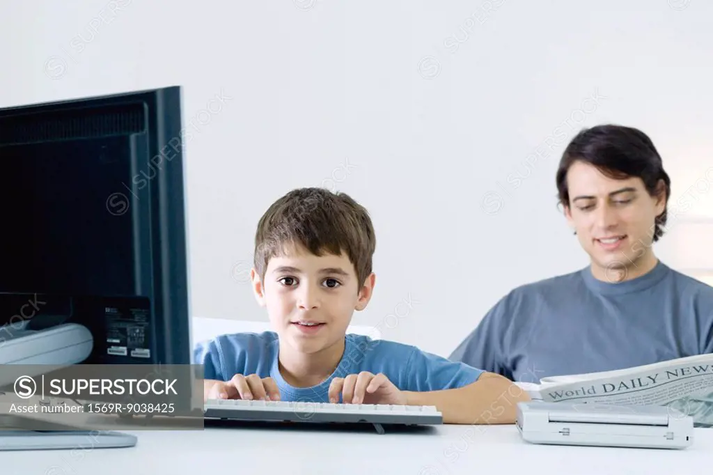 Little boy using computer, smiling at camera, father reading newspaper in background