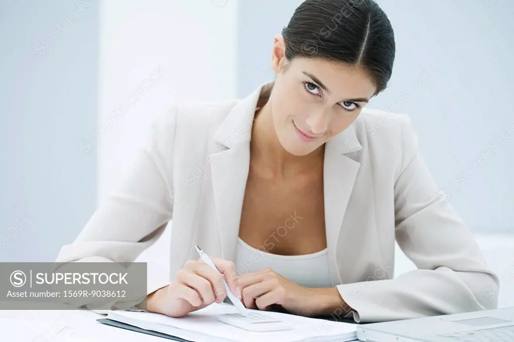 Young businesswoman using pen to enter numbers on calculator, looking at camera