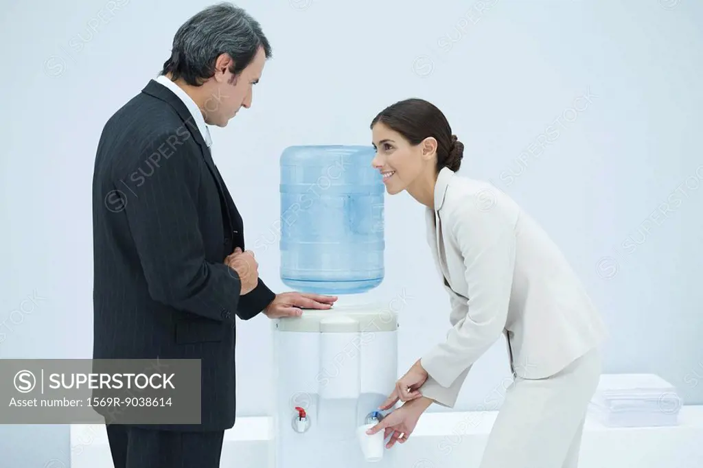Two professionals chatting beside water cooler, woman filling disposable cup