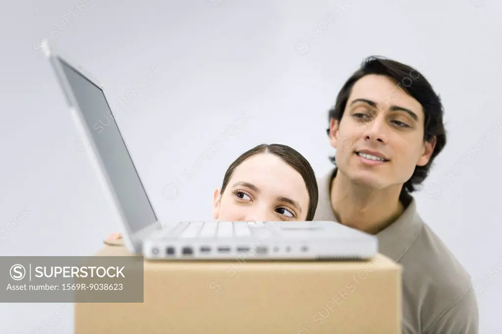 Man and woman peeking at laptop computer sitting on top of cardboard box