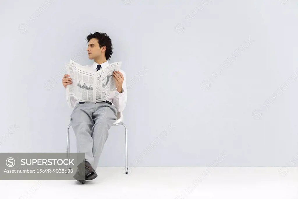 Businessman sitting in chair, holding newspaper, looking away