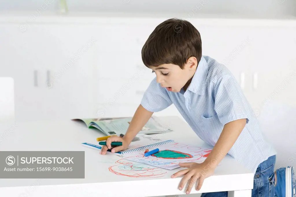 Boy leaning on table, holding crayon, colorful drawing on paper and table