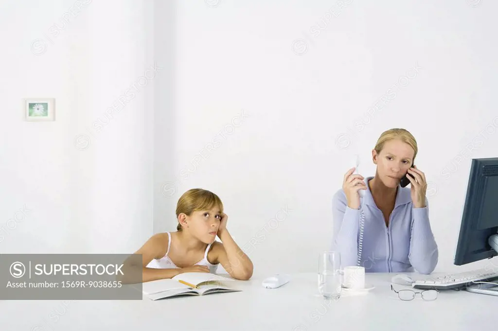 Woman sitting in front of computer, using cell phone and landline phone, daughter sulking nearby
