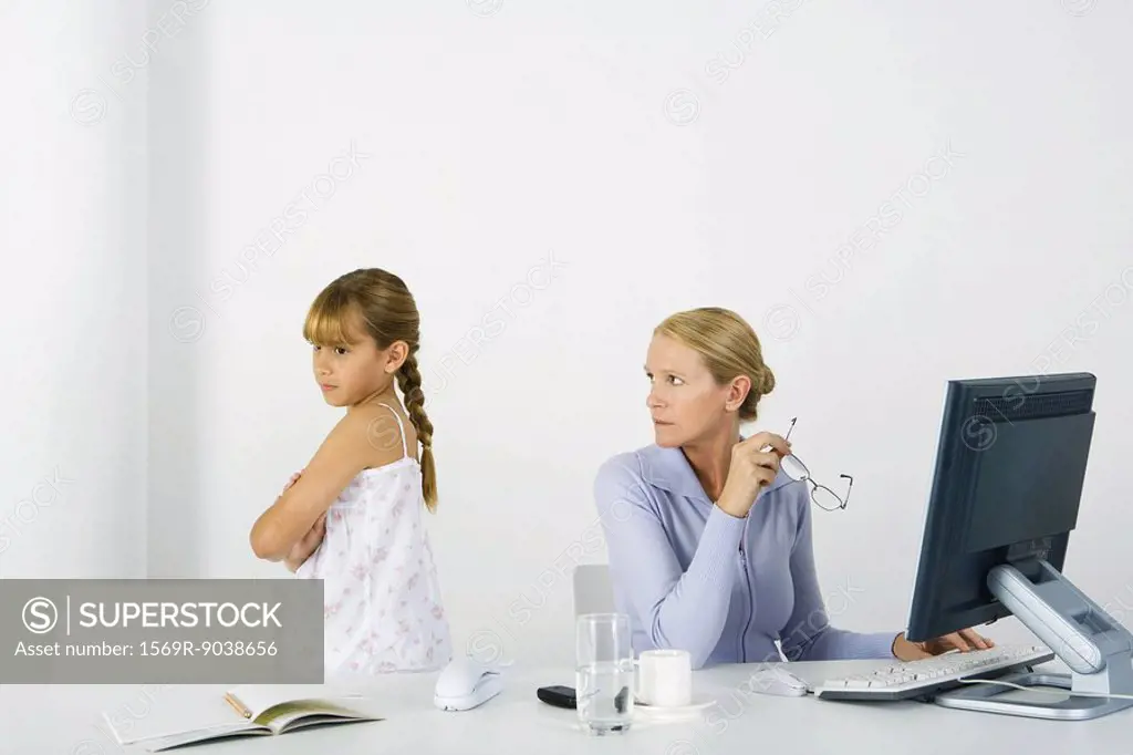 Woman sitting before computer, looking over shoulder at daughter sulking nearby