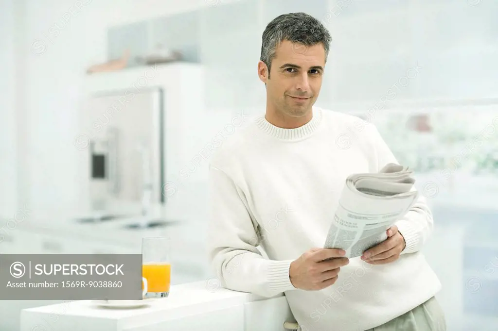 Man leaning against kitchen counter, holding newspaper, smiling at camera