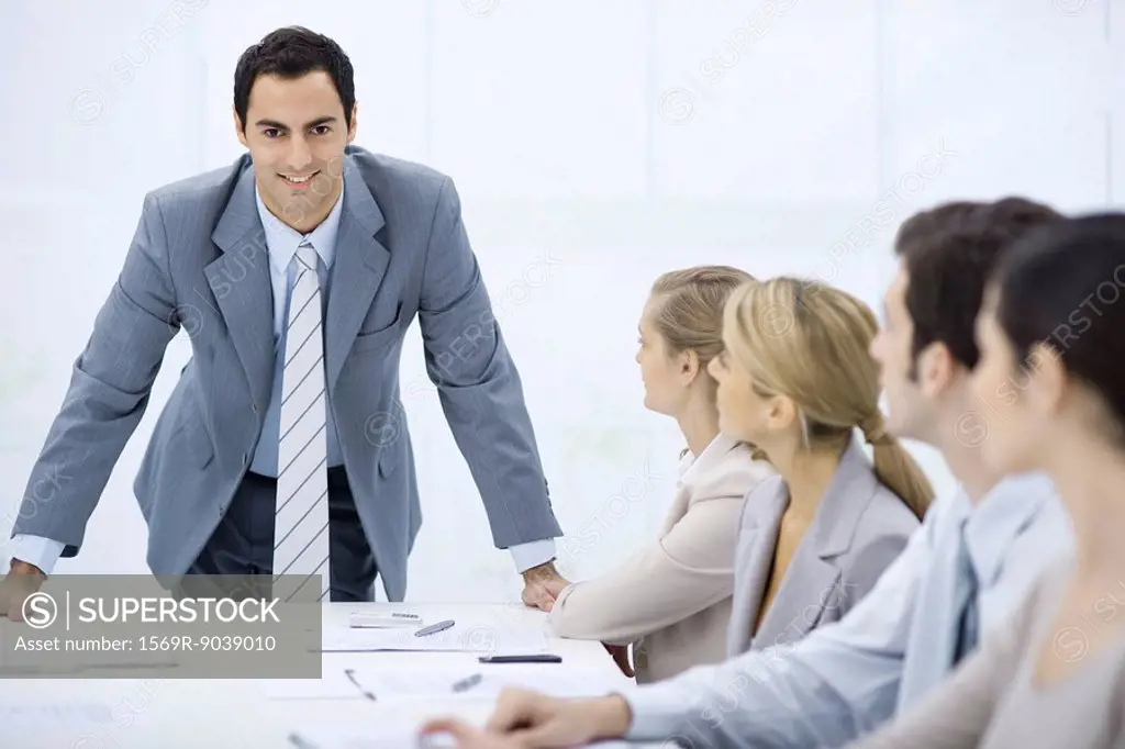 Businessman leaning over table addressing colleagues, smiling at camera