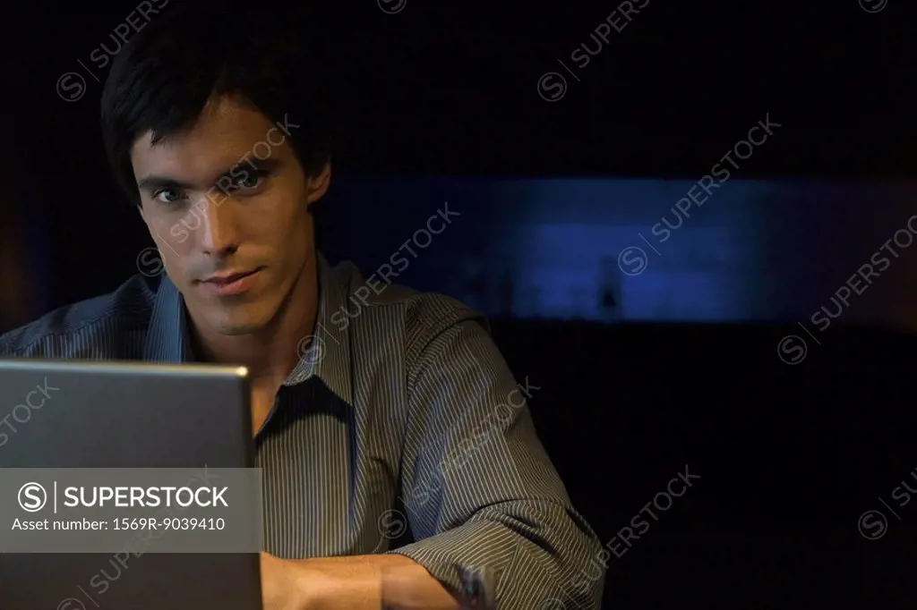 Man sitting in darkly lit room at table with laptop, looking at camera, portrait