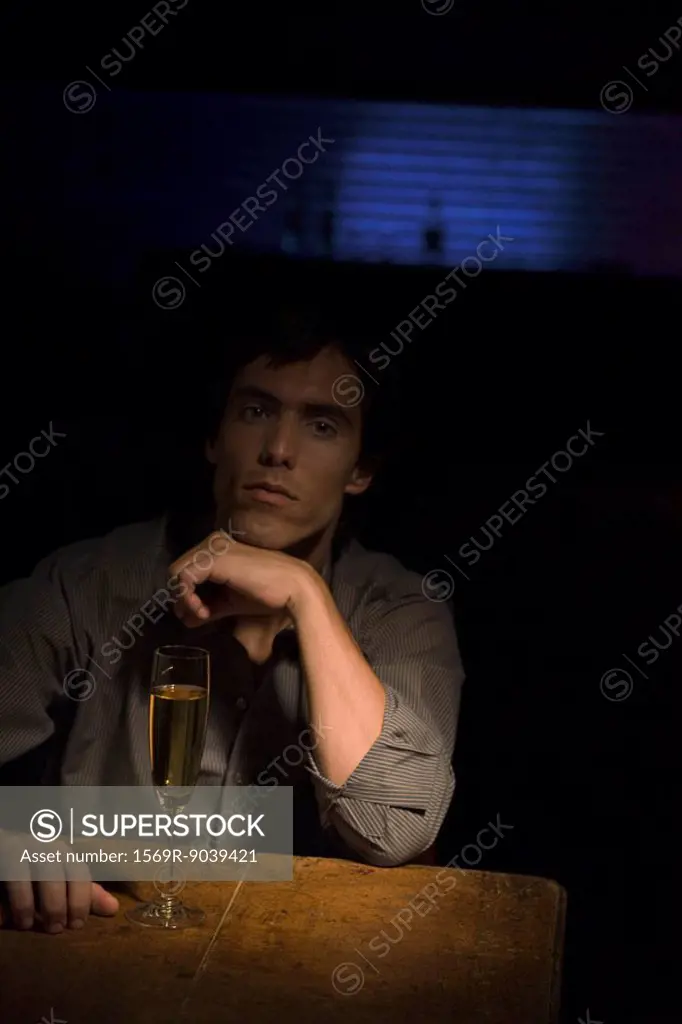 Man sitting in the dark, hand under chin, spotlight on glass of champagne on table